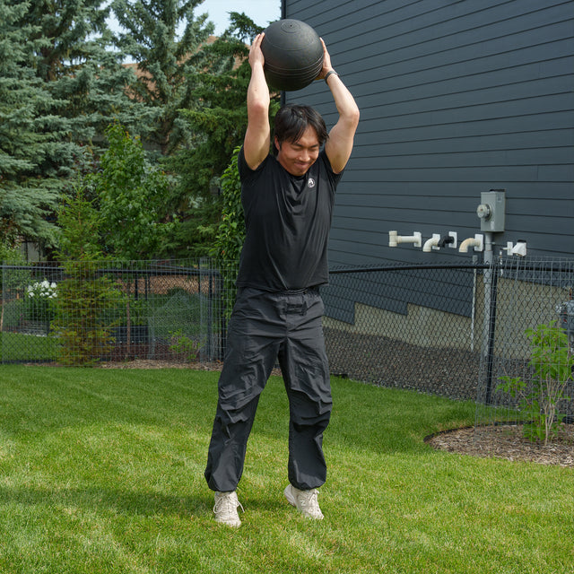 Dressed in black workout gear, an athlete lifts a Bells of Steel Slam Ball overhead while standing on backyard grass, preparing to slam it down—a dynamic way to unleash explosive power and ramp up the intensity of your cardio workouts.