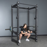 A woman does squats in a gym power rack using the Bells of Steel Safety Squat Bar - The SS4, with weight plates on her shoulders. She wears a black T-shirt, black shorts, and white sneakers.
