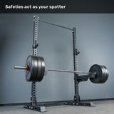 A loaded barbell rests on the safeties of a Bells of Steel Manticore Squat Stand Builder (3" x 3", 1" Holes) in an empty gym, with text above stating, "Safeties act as your spotter.