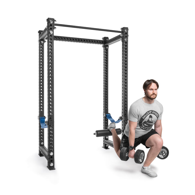 A man performs Bulgarian split squats using a Bells of Steel Split Squat Leg Roller Rack Attachment. Wearing a gray T-shirt and black shorts, he holds a dumbbell in each hand while his back leg rests on the attachment for support.