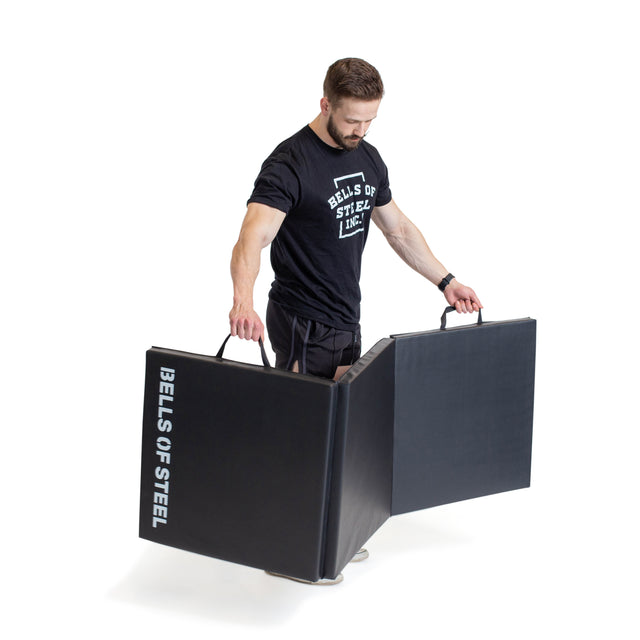 A man in a "Bells of Steel" shirt holds and opens a large black Trifold Mat with handles and the "Bells of Steel" logo on the side, set against a plain white background.