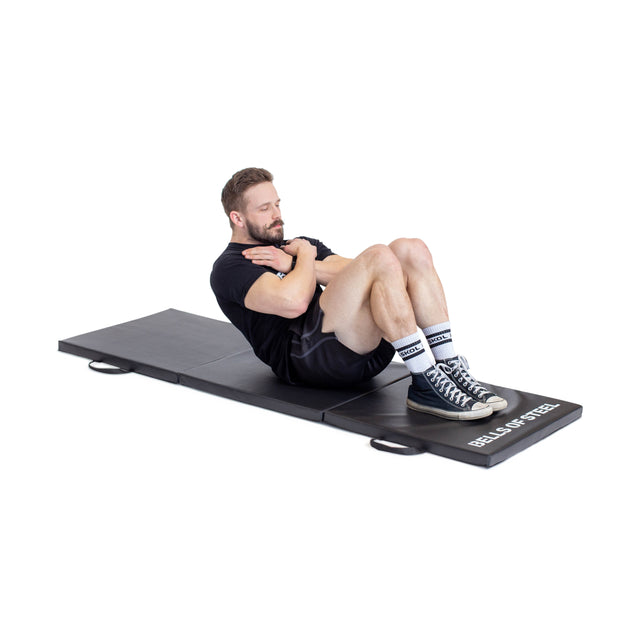 A man in athletic wear does crunches on a black Bells of Steel Trifold Mat, featuring a non-slip surface and visible branding. His arms are crossed over his chest and his knees are bent.