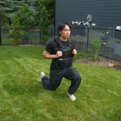 A person wearing the Bells of Steel Weighted Vest and Plates along with black workout clothes performs a lunge on a green lawn in a fenced backyard.