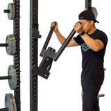 A person in a black shirt and cap adjusts the Bells of Steel Y Dip Bar Rack Attachment on the gym's heavy-duty rack, surrounded by weight plates. UHMW lining protects both the attachment and metal rack from wear.