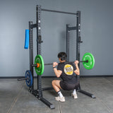 A person wearing a black shirt and shorts performs a barbell squat in a Bells of Steel Hydra Half Rack - Prebuilt (3" x 3", ⅝" Holes) in a gym, holding a barbell with green and blue plates across their upper back.