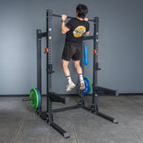 A person in a black shirt and shorts does a pull-up on the Bells of Steel Hydra Half Rack - Prebuilt (3" x 3", ⅝" Holes) in a home gym, with weights on the frame and a gray wall behind.