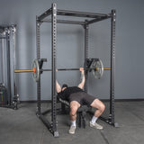 A person performs a bench press on a bench inside a power rack, using the Bells of Steel Monolift Rack Attachment to hold the barbell above their chest.