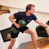 A man in a navy T-shirt and shorts works out indoors, performing a bent-over row with the Nuobell NÜOBELL Adjustable Dumbbells in a modern room with wooden floors and a fireplace. He looks focused and determined.