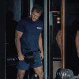 A man in a blue t-shirt and shorts lifts a Bells of Steel NÜOBELL-S Adjustable Dumbbell in the gym, standing near a mirror with a focused expression.