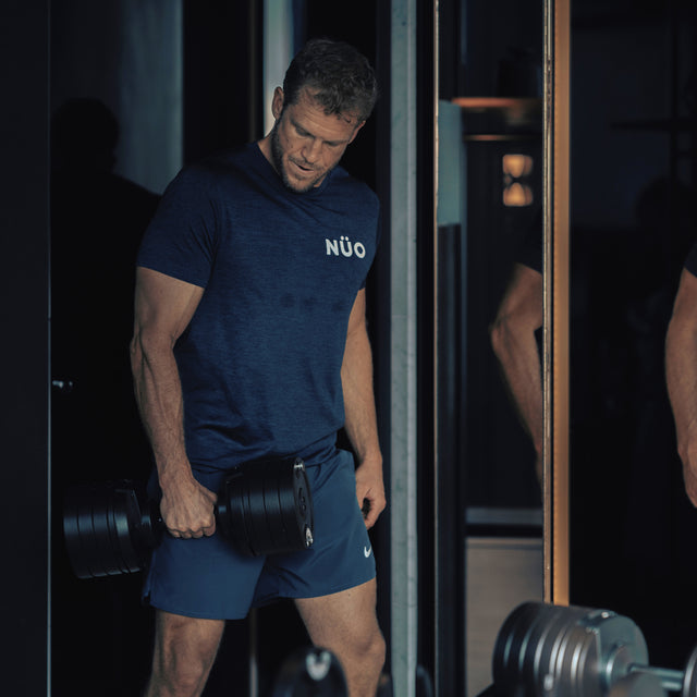 A muscular man in a blue NUÖ shirt and shorts stands in a dimly lit gym, holding a Bells of Steel NÜOBELL-S Adjustable Dumbbell by his side and looking down with focus. Gym equipment appears in the background.