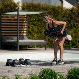 A woman in athletic wear does a bent-over row outdoors near a pool, using Nuobell NÜOBELL Adjustable Dumbbells with medium-knurled handles. Machined steel weight plates are on the wooden deck, surrounded by lush greenery.