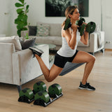 A woman in athletic wear does a split squat with Nuobell NÜOBELL Adjustable Dumbbells, one foot on a sofa, while extra machined steel weight plates rest on the floor of her modern living room.