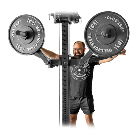 A man in a black t-shirt and shorts smiles behind the Bells of Steel Shoulder Boulder/Chest Fly Attachment, ready for a chest fly workout, with large branded weight plates beside the power rack.