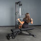 A woman with long brown hair performs a lat pulldown on the Bells of Steel Cable Tower, using the weight stack while seated on a workout bench in a gym with gray walls and flooring.