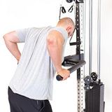 A person in a grey shirt uses the Bells of Steel Cable Tower for triceps exercises, gripping a horizontal bar on the weight stack with both hands and leaning forward during the workout.