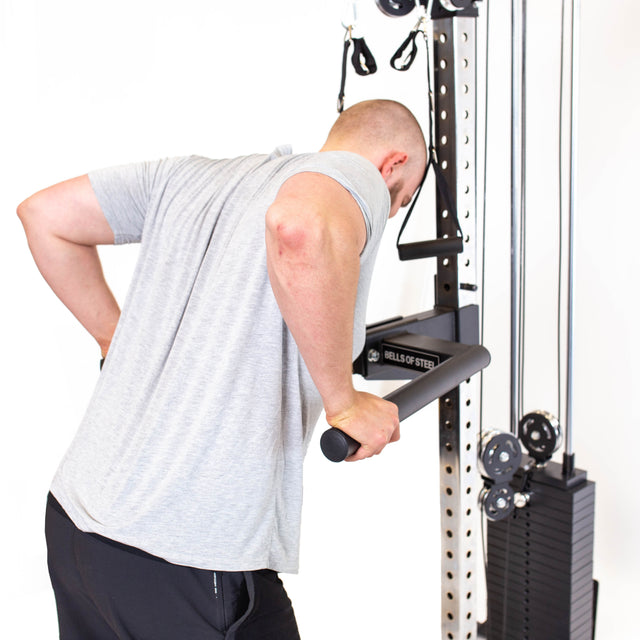 A person in a grey shirt uses the Bells of Steel Cable Tower for triceps exercises, gripping a horizontal bar on the weight stack with both hands and leaning forward during the workout.