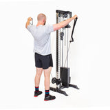 A person in a gray t-shirt and black shorts uses the Bells of Steel Cable Tower, standing with arms raised and pulling adjustable handles outward for a strength training exercise.