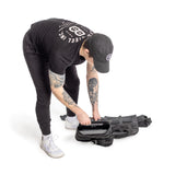 Dressed in black athletic gear and a cap, an individual with visible arm tattoos places a Bells of Steel Weighted Vest and Plates into a black backpack against a white background.
