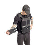 A man in black attire adjusts the strap of the Bells of Steel Weighted Vest and Plates, featuring an adjustable fit, with the brand’s logo displayed on front against a plain white background.