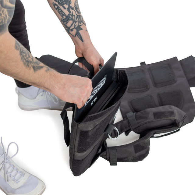 A person with tattooed arms adds a metal plate to the Bells of Steel Weighted Vest and Plates, shown open on a white background. This workout accessory offers an adjustable fit. Only their lower body and hands are in view.