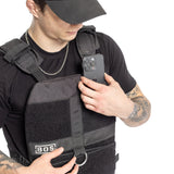 A tattooed individual wearing a black Bells of Steel Weighted Vest, designed for training plates and featuring a logo and metal ring on the front, adjusts the pocket while holding a smartphone. They complete their look with a chain necklace and cap.